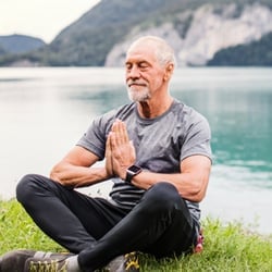 Senior bearded man in yoga meditation pose sitting over green grass by lake and mountains.