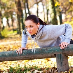 A woman in sportswear and listening to music with earphones while doing push-ups over a wooden ledge in a park.